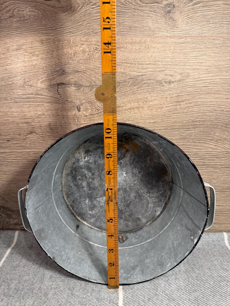 Op de afbeelding: Een ronde gegalvaniseerde metalen kom met twee handgrepen aan de zijkant en een donkere rand. Een gele opvouwbare houten liniaal ligt in het midden en toont een diameter van ongeveer 26,5 cm. De binnenkant vertoont sporen van slijtage.