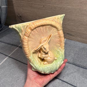 Könnte beinhalten: Eine Keramikvase mit einem Relief eines Kaninchens in einer grasbewachsenen Umgebung. Die Vase ist hellbeige mit grünen und braunen Akzenten. Das Kaninchen ist hellbeige und hat lange Ohren. Die Vase wird in einer Hand gehalten.