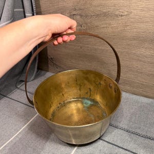 May include: A round brass bucket with a weathered patina and visible green oxidation spots on the interior base. The vessel features a thin, dark metal swing handle attached to the sides with two rivets, creating a simple, functional design.