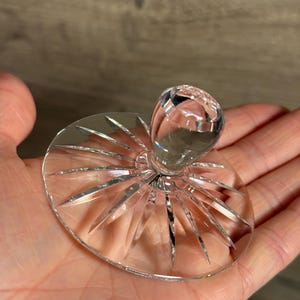 Op de afbeelding: Een heldere kristallen glazen flesstopper met een ronde, facetgeslepen knop en een cirkelvormige basis. De basis heeft een sterrenpatroon met puntige randen. De stopper wordt in een hand gehouden.