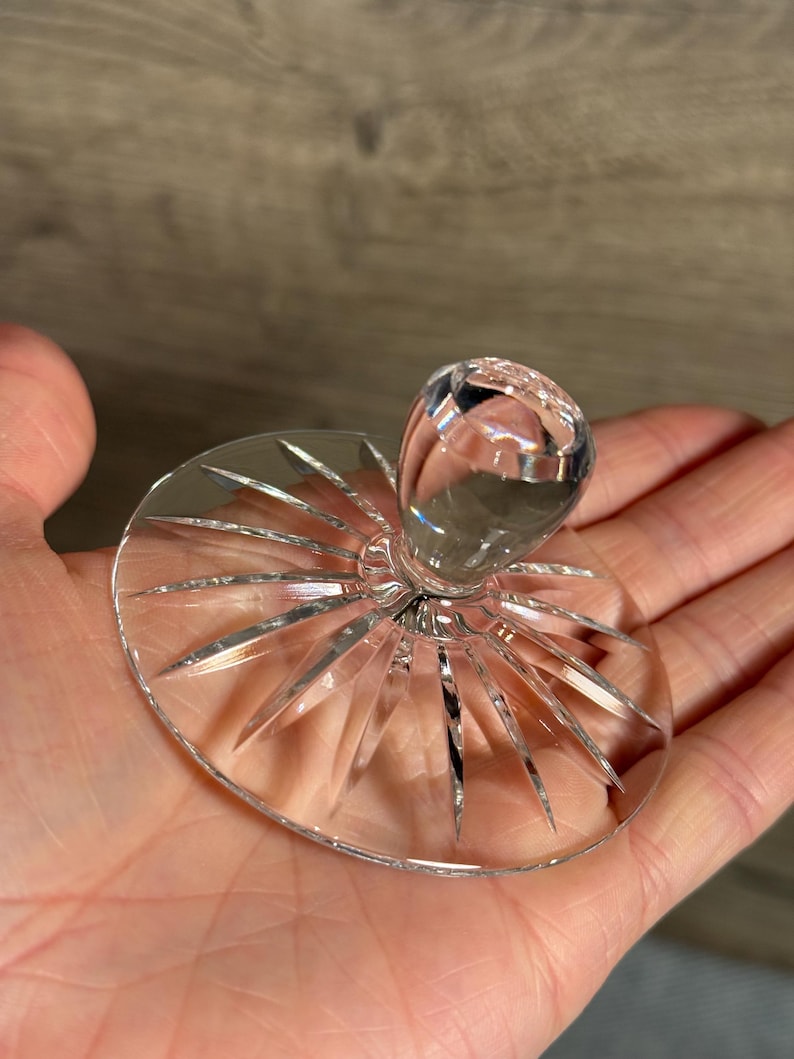 Op de afbeelding: Een heldere kristallen glazen flesstopper met een ronde, facetgeslepen knop en een cirkelvormige basis. De basis heeft een sterrenpatroon met puntige randen. De stopper wordt in een hand gehouden.