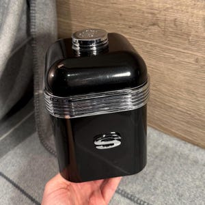 Op de afbeelding: Een zwarte, rechthoekige container met een glanzend, reflecterend oppervlak. De container heeft een zilverkleurige band in het midden en een zilveren dop bovenop. Een zilveren "S"-logo staat op de voorkant.