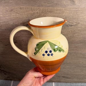 Puede incluir: Una jarra de cerámica con la parte superior de color crema y la base de color naranja sólido. El frente está decorado con un diseño pintado a mano de hojas verdes y pequeñas bayas de color azul oscuro. Tiene un borde ancho y un asa.