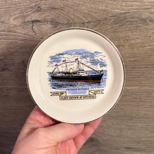 Könnte beinhalten: Kleine, runde, weiße Platte mit goldenem Rand. Die Platte zeigt eine detaillierte Illustration eines Schiffs mit Flaggen, vor blauem Himmel und Meer. Text: "H.M. Yacht Britannia", "June 28" und "Fleet Review at Spithead 1977".
