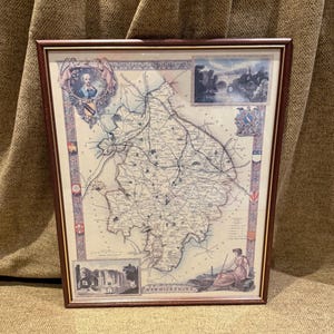 May include: Framed vintage map of Warwickshire, England, with detailed cartography. The map features illustrations of historical figures, buildings, and landscapes. The frame is a dark brown color.