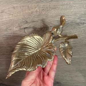 May include: A brass decorative piece featuring a bird perched on a branch with two leaves. The large leaf is in the foreground, and the bird is facing right. The piece is held in a hand against a wooden background.