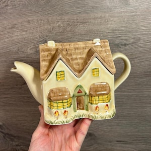 Puede incluir: Una tetera de cerámica con forma de casa, con tejado marrón y paredes color crema. La casa tiene dos ventanas, una puerta y una chimenea. La tetera tiene un pico curvo y un asa. El diseño incluye detalles en amarillo y verde.