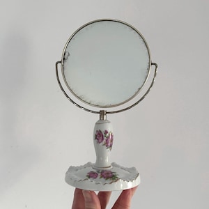 May include: A vintage vanity mirror with a round, silver-toned frame and a white porcelain base. The base and handle are decorated with pink floral patterns. The mirror is held in place by a curved metal support.