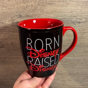 Peut inclure: Mug &agrave; caf&eacute; noir avec int&eacute;rieur et anse rouges. Le mug porte l'inscription "BORN Disney RAISED Disney" en lettres blanches et rouges. Un cadeau parfait pour les fans de Disney.