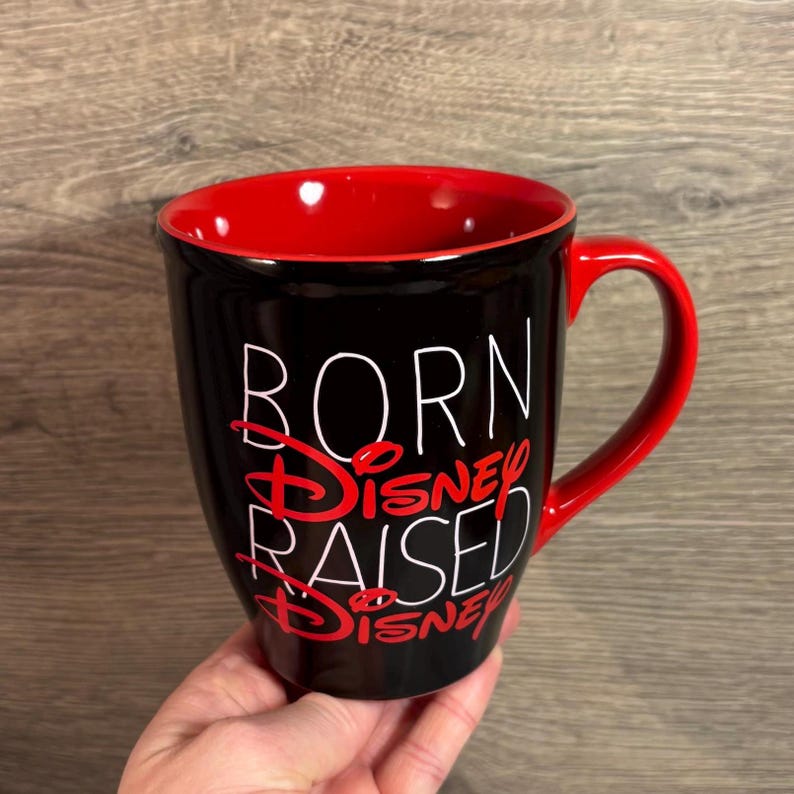 Peut inclure: Mug &agrave; caf&eacute; noir avec int&eacute;rieur et anse rouges. Le mug porte l'inscription "BORN Disney RAISED Disney" en lettres blanches et rouges. Un cadeau parfait pour les fans de Disney.