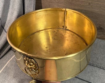 May include: A round, antique brass planter with a lion's head ring handle and decorative foot. The interior and exterior of the planter have a golden hue. The planter is likely used for decorative purposes.