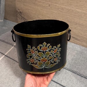 May include: A black oval metal bucket featuring a hand-painted floral design in shades of orange, green, and white. The bucket has a gold-painted rim, two side ring handles, and a black interior. The exterior surface is smooth and glossy.