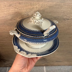May include: A white porcelain soup tureen with a blue and gold floral design. The tureen has a lid with a knob and a matching serving spoon.