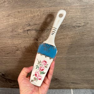 以下が含まれることがあります： 白いと青いセラミックケーキサーバー、バラの花柄。 サーバーは、金色のアクセントと金色の透かし模様の装飾的な青い部分を備えたハンドルを備えています。 ピンクのバラと緑の葉がサーバーの下部を飾っています。