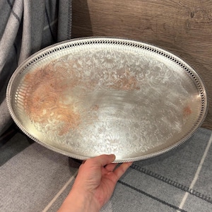 Könnte beinhalten: Ovale silberne Servierplatte mit aufwendigem Blumen- und Scroll-Design. Das Tablett hat einen dekorativen durchbrochenen Rand. Das Tablett wird von einer Hand gehalten. Das Tablett liegt auf einer grau-weiß karierten Decke.