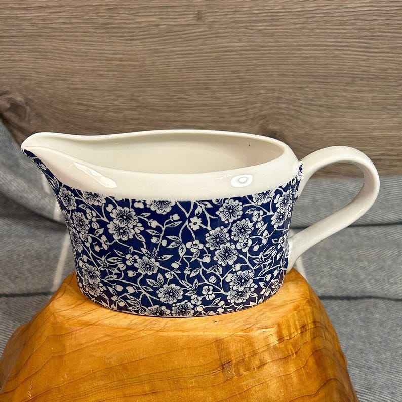 May include: A white ceramic gravy boat with a blue and white floral pattern. The boat has a handle and a spout.