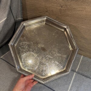 May include: Silver-coloured, octagonal serving tray with ornate floral and scrollwork detailing. The tray has a decorative, beaded edge and a pierced gallery. The tray is resting on a grey and white striped surface.