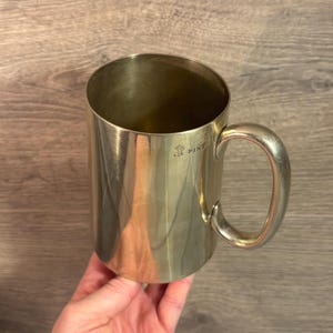 Könnte beinhalten: Ein silberfarbener Pint-Becher mit einem großen Henkel. Der Becher hat eine zylindrische Form und eine glatte Oberfläche. Das Wort "PINT" ist auf der Seite eingeprägt. Der Becher wird von einer Person gehalten.