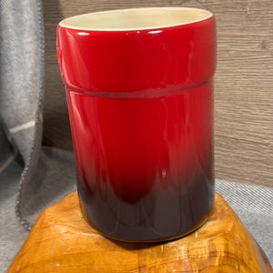 May include: Red and black ceramic utensil holder with a rounded shape.