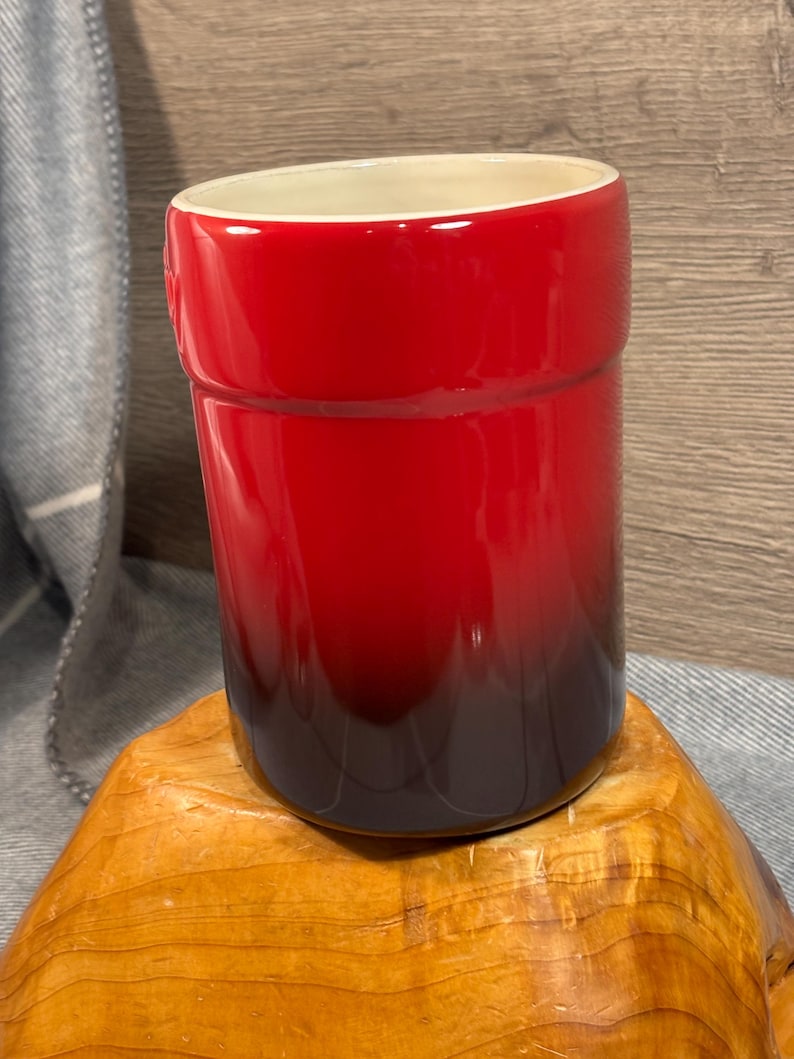 May include: Red and black ceramic utensil holder with a rounded shape.