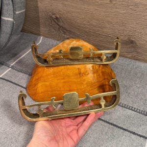 Peut inclure: Deux traîneaux décoratifs en laiton avec patins incurvés et détails ornés. Les traîneaux sont tenus au-dessus d'un bol en bois avec une finition naturelle. L'arrière-plan comprend un tissu rayé gris et blanc.
