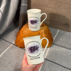 May include: Two white ceramic mugs with a green rim. Both mugs feature a black and white image of a steam engine with the text "Boss-on-Wye Steam Engine Society" and "44th Annual Rally Welland 2008".