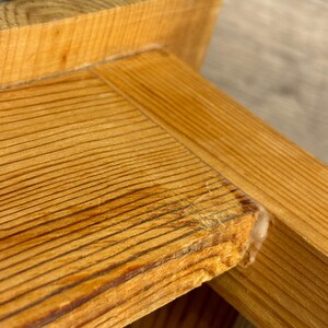 May include: Close-up of a wooden furniture corner. The wood has a natural, light brown color with visible grain patterns. The corner shows a joint with glue residue, highlighting the construction details. The overall aesthetic is rustic and natural.