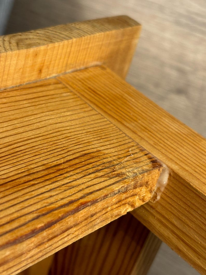 May include: Close-up of a wooden furniture corner. The wood has a natural, light brown color with visible grain patterns. The corner shows a joint with glue residue, highlighting the construction details. The overall aesthetic is rustic and natural.