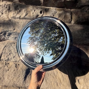 May include: A circular silver-toned metal tray with a polished, reflective surface. The tray features a smooth, raised rim and a wide, flat border. The reflective centre displays a clear view of trees and a bright sun against a blue sky.