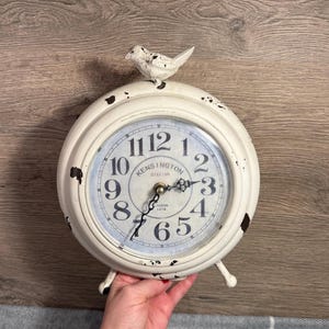 May include: Cream-coloured vintage-style clock with black numerals and hands. The clock face reads "Kensington Station London 1879." A small, decorative bird sits atop the clock. The clock is held by a hand.