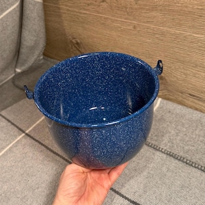 Peut inclure: Un bol en émail bleu avec des mouchetures blanches. Le bol a deux petites poignées et une forme arrondie. Le bol est posé sur une surface à motifs gris et blancs.