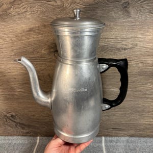 May include: A silver aluminum coffee pot with a black handle and a lid. The pot has a label that reads "Trellis".