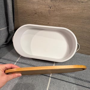 May include: An oval-shaped white storage container with a smooth interior and small side handles. A separate, matching oval-shaped wooden lid is held horizontally in the foreground, showing its natural light brown wood grain and smooth finish.