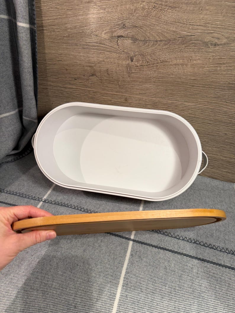 May include: An oval-shaped white storage container with a smooth interior and small side handles. A separate, matching oval-shaped wooden lid is held horizontally in the foreground, showing its natural light brown wood grain and smooth finish.