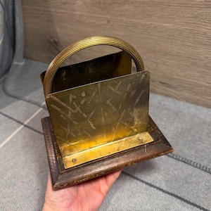 May include: A brass napkin holder featuring a rectangular metal plate with a textured, scratched surface finish and a rounded, ribbed brass handle. The metal component is securely mounted onto a dark, stained wooden base with beveled edges.