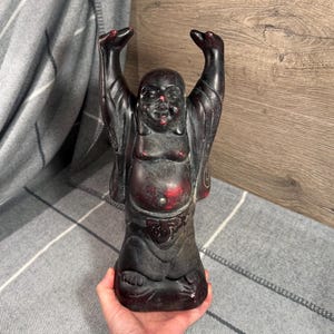 Puede incluir: Estatua oscura de Buda sentado con los brazos levantados. La estatua tiene un acabado negro con detalles en rojo en el pecho y la cara. La figura está sentada y tiene una expresión serena.