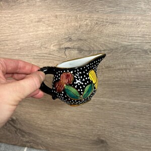 May include: A small black ceramic creamer with a white and gold polka dot pattern. The creamer has a floral design with red, yellow, and green accents.