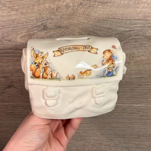 Puede incluir: Hucha de cerámica color crema con forma de bolso, con una ilustración caprichosa de animales del bosque y un oso de peluche. El banco tiene una ranura para monedas y el texto "Rambling Ted" en una pancarta.