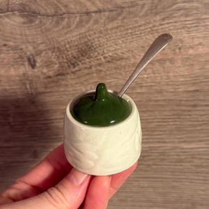 Puede incluir: Un pequeño azucarero de cerámica blanquecina con tapa verde oscuro y una cucharita plateada. El cuenco tiene un esmalte craquelado y un sutil diseño floral en relieve. La tapa tiene un pequeño asa redondeada.