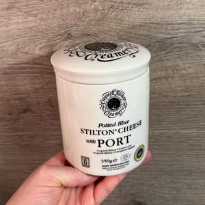 Op de afbeelding: Een witte keramische pot met een deksel, met het opschrift "Potted Blue Stilton Cheese with Port". De pot heeft zwarte tekst en een decoratief embleem. De pot bevat 350g kaas.