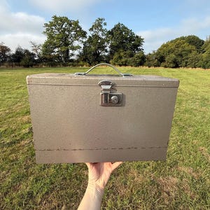Puede incluir: Una caja de almacenamiento de metal vintage con asa y pestillo. La caja es de color gris y parece estar hecha de metal. El asa y el pestillo son plateados. La caja se sostiene frente a un campo de hierba con árboles al fondo.