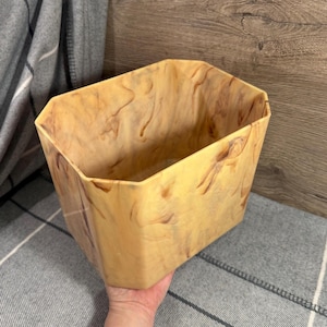 May include: A rectangular, open-top container with a marbled pattern in shades of tan and brown. The container has a geometric shape with angled sides and a wide opening. The material appears to be a solid, possibly plastic or resin.