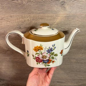 Könnte beinhalten: Eine weiße Keramik-Teekanne mit goldenem Rand und Deckel. Die Teekanne ist mit einem Blumenmuster mit orangefarbenen, gelben, rosa und blauen Blumen verziert. Der Griff und der Ausguss sind weiß mit schwarzem Rand.