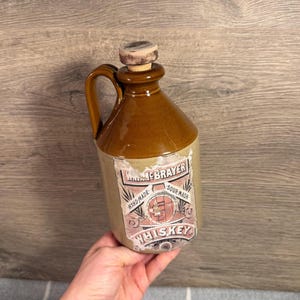 Puede incluir: Una jarra de whisky de cerámica marrón con asa y tapón de corcho. El frente presenta una etiqueta de papel desgastada con el texto W.H. McBRAYER, HAND-MADE, SOUR MASH y WHISKEY, junto con un gráfico central de un barril y equipo de destilería.