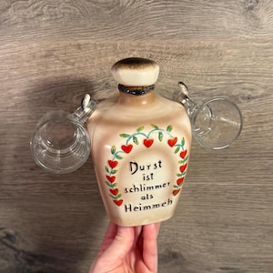 Könnte beinhalten: Eine beige Keramikflasche mit braunem Verschluss und zwei daran befestigten Gläsern. Die Flasche ist mit einem Kranz aus roten Herzen und grünen Blättern verziert, und der Text "Durst ist schlimmer als Heimweh" ist auf der Vorderseite geschrieben.