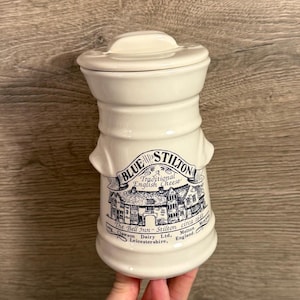May include: A white ceramic jar with a lid, featuring a blue and white design. The jar is labeled "BLUE STILTON Traditional English Cheese" with an illustration of a building and text. The jar is held by a hand.