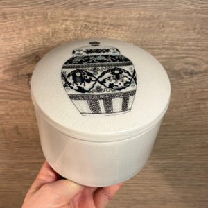 May include: White ceramic jar with a lid featuring a blue and black design of a traditional Chinese vase with floral and geometric patterns.