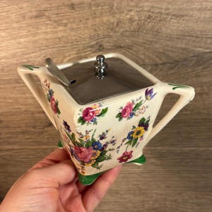 May include: A vintage floral patterned sugar bowl with a silver lid and a spoon. The bowl has a square shape with rounded edges and green accents.