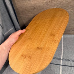 May include: An oval-shaped wooden serving tray with a smooth, light brown finish and visible natural wood grain patterns. The tray features rounded edges and a flat surface, showcasing the organic texture and warm tones of the wood material.