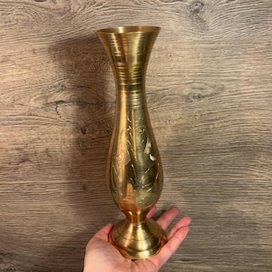 May include: A vintage brass vase with a flared top and an etched design. The vase has a round base and a slender body. The brass has a warm, golden hue. The vase is held in a hand. The vase is approximately 20 cm tall.